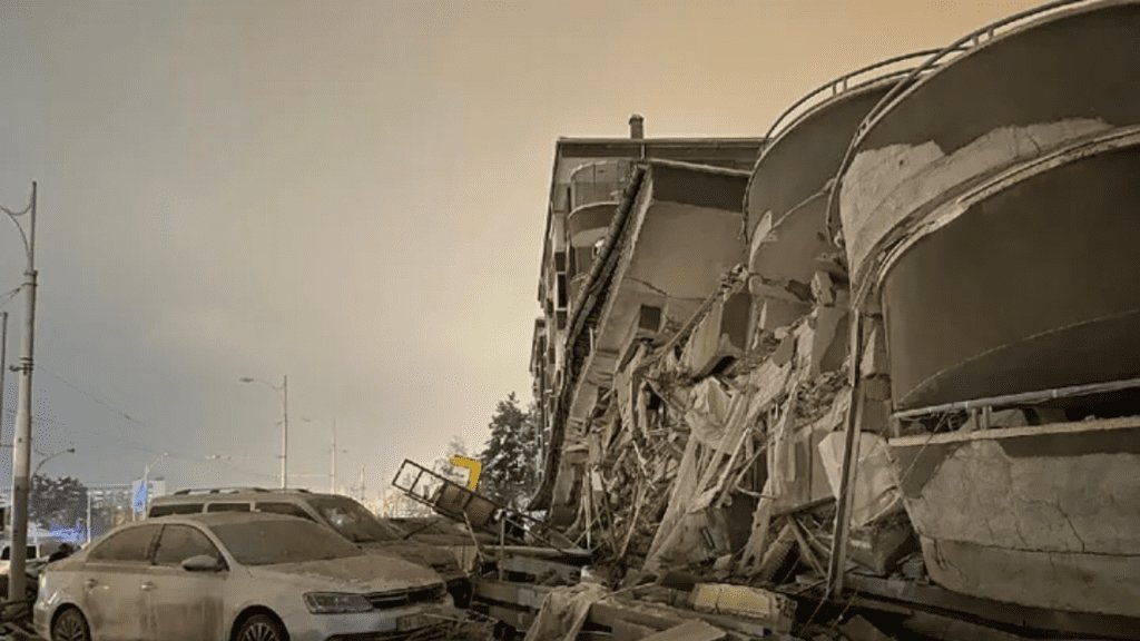 Damaged vehicles sit parked in front of a collapsed building.